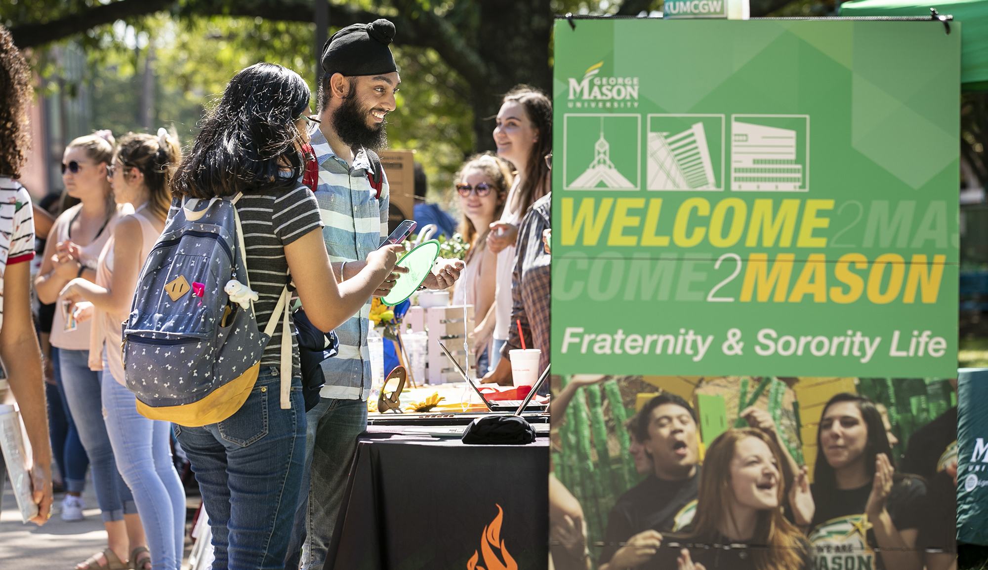 Group of smiling students standing around a table. A sign in the front of the table says "WELCOME2MASON Fraternity & Sorority Life"